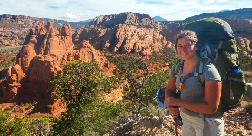 A person wearing a backpack smiles in front of a desert landscape with large red rock formations.